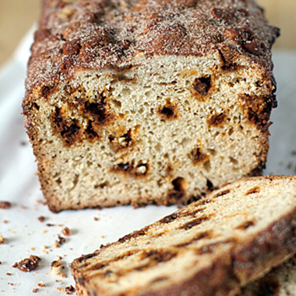 A loaf of cinnamon swirl bread with a sugary, cinnamon-topped crust, partially sliced, resting on parchment paper with crumbs scattered around.