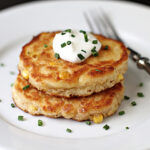 Two golden corn fritters stacked on a white plate, topped with a dollop of sour cream and sprinkled with chopped chives. A fork rests beside the fritters.