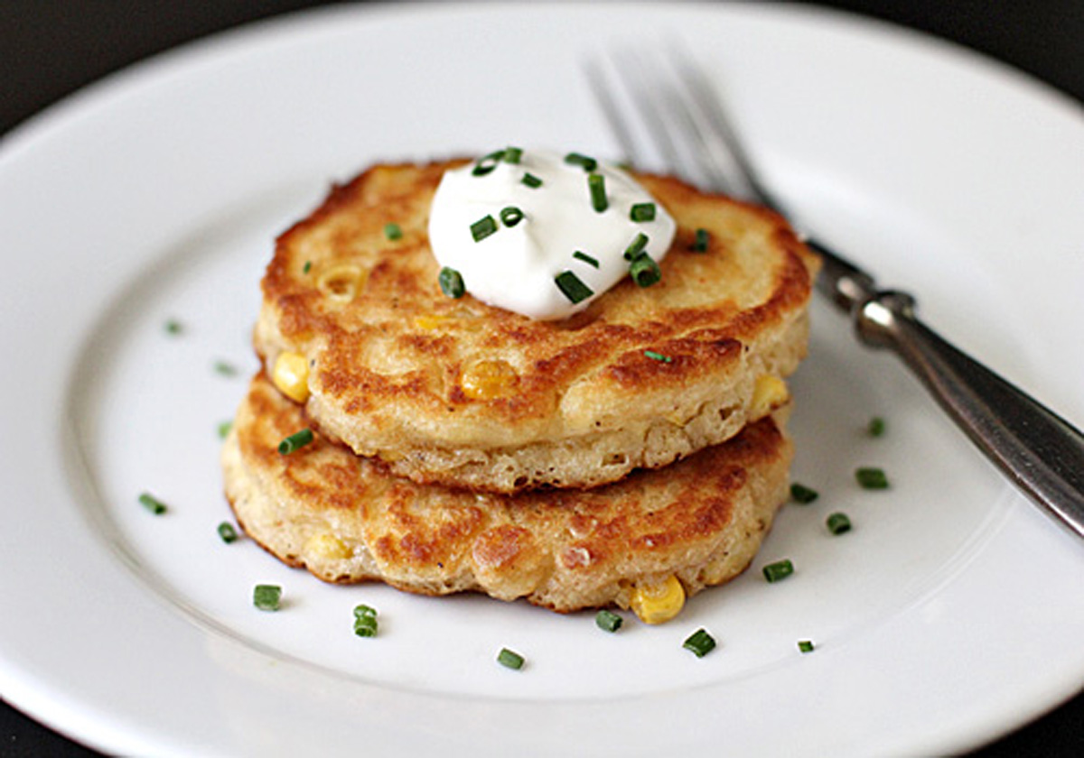Two golden corn fritters stacked on a white plate, topped with a dollop of sour cream and sprinkled with chopped chives. A fork rests beside the fritters.
