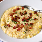 A white bowl filled with creamy grits topped with crispy bacon bits and chopped herbs, with a silver spoon resting on the side.