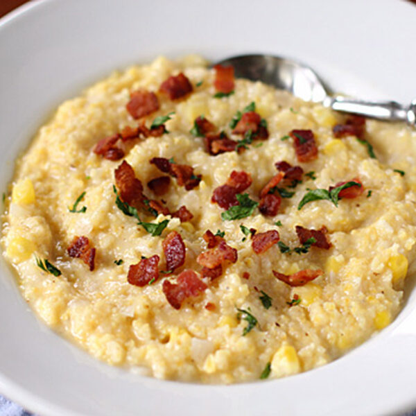 A white bowl filled with creamy grits topped with crispy bacon bits and chopped herbs, with a silver spoon resting on the side.