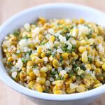 A white bowl filled with sautéed corn kernels topped with chopped herbs and crumbled cheese, with lime wedges blurred in the background.