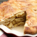A baked apple pie with a golden, flaky crust sits in a pie dish. A large slice has been removed, revealing layers of cooked apple filling inside.