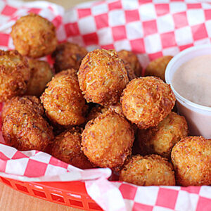 A red basket lined with red-and-white checkered paper holds a pile of golden-brown hush puppies next to a small cup of creamy dipping sauce.