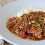 A plate of smothered steak in tomato gravy, garnished with chopped green onions, served alongside a mound of creamy mashed potatoes. A fork and knife rest on a folded napkin beside the plate.