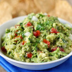 A white bowl filled with chunky guacamole, containing diced tomatoes, onions, and cilantro, sits on a blue cloth with tortilla chips visible in the background.
