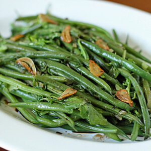 A white plate filled with cooked green beans, garnished with sautéed garlic slices and herbs. The dish appears fresh and vibrant, with a glossy finish from a light seasoning or oil.