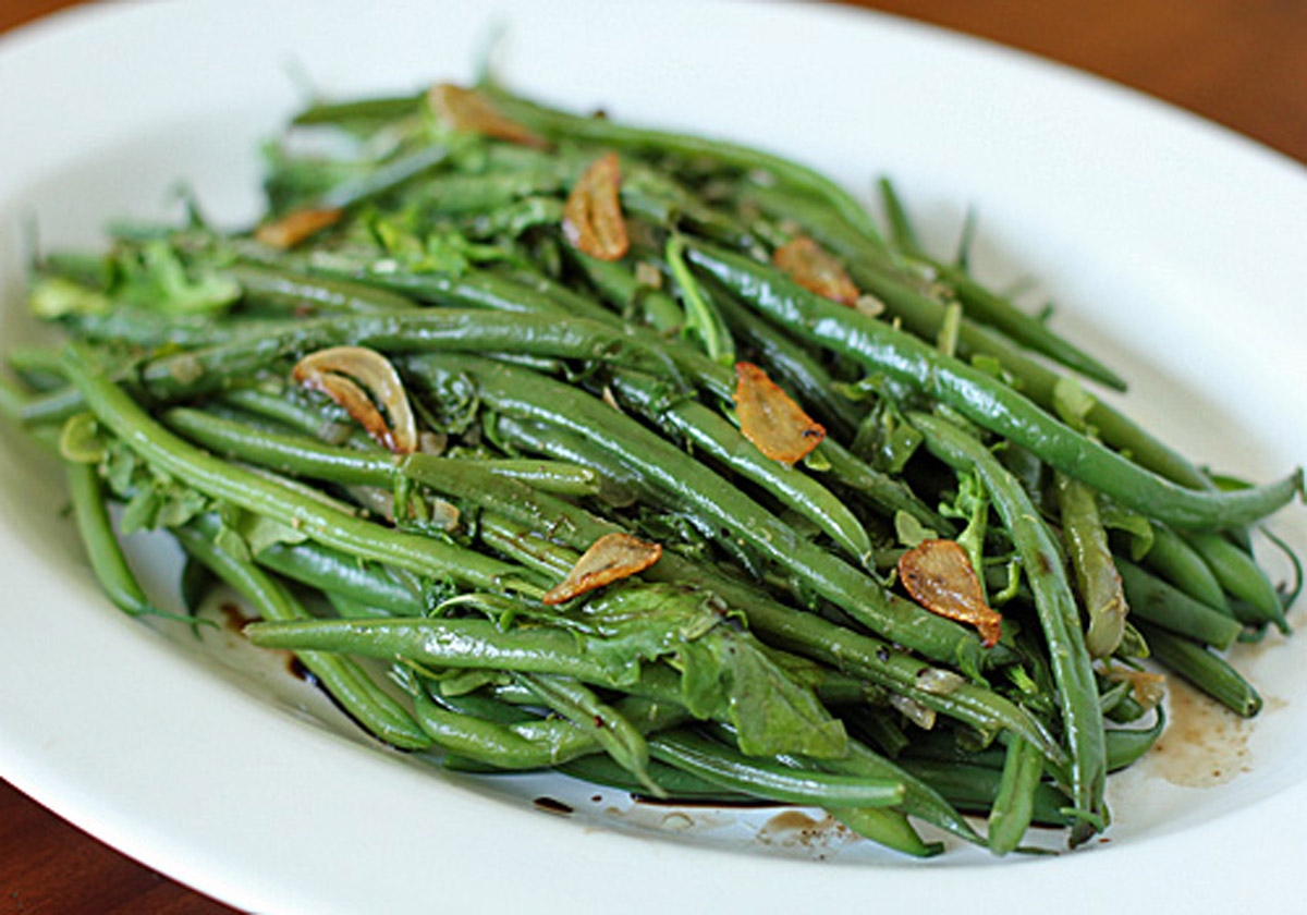 A white plate filled with cooked green beans, garnished with saut&eacute;ed garlic slices and herbs. The dish appears fresh and vibrant, with a glossy finish from a light seasoning or oil.