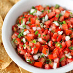 A white bowl filled with fresh pico de gallo made of diced tomatoes, onions, cilantro, and jalapeños, next to a pile of tortilla chips.