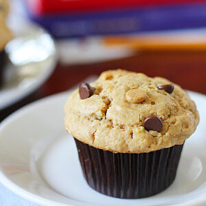 A freshly baked chocolate chip muffin in a brown paper liner sits on a white plate, with a blurry background featuring a glass of milk and colorful books.