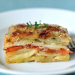 A close-up of a neatly cut slice of potato and tomato gratin, garnished with herbs, on a white plate with a fork resting beside it. The layers of potatoes and tomatoes are clearly visible.