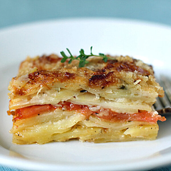 A close-up of a neatly cut slice of potato and tomato gratin, garnished with herbs, on a white plate with a fork resting beside it. The layers of potatoes and tomatoes are clearly visible.