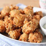 A white plate filled with golden-brown hush puppies, served next to a small bowl of creamy dipping sauce. A bottle of hot sauce is visible in the background.
