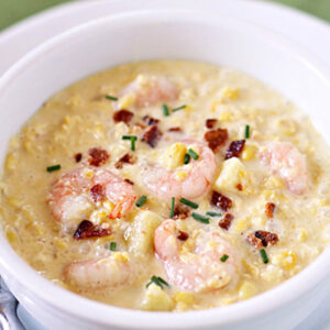 A white bowl filled with creamy shrimp chowder, garnished with crumbled bacon and chopped chives, sits next to a silver spoon on a white plate.