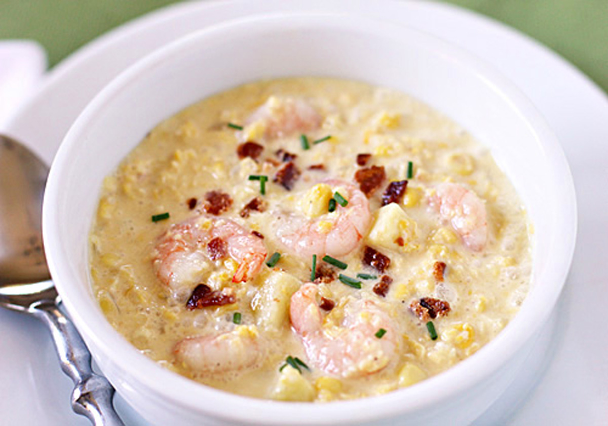 A white bowl filled with creamy shrimp chowder, garnished with crumbled bacon and chopped chives, sits next to a silver spoon on a white plate.
