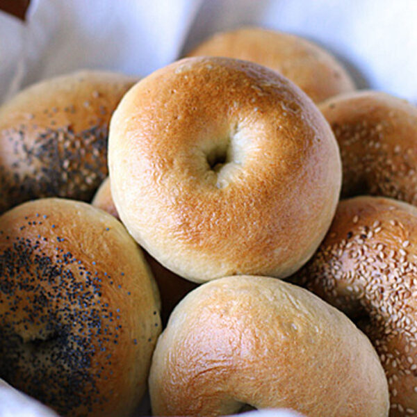 A basket lined with white cloth holds several plain, poppy seed, and sesame seed bagels. The bagels are golden brown and arranged closely together.