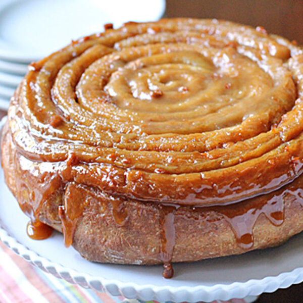 A large glazed cinnamon roll cake sits on a white platter, with caramel sauce dripping down the sides. Stacked plates and forks are in the background on a colorful cloth.
