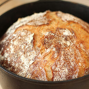 A round, golden-brown loaf of crusty bread with a rustic, cracked surface sits in a black cast iron pot.