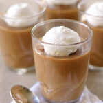 Four glasses of creamy caramel pudding topped with dollops of whipped cream, arranged on a light surface with a silver spoon and a white napkin in front.