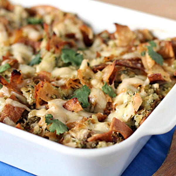 A baked casserole in a white dish, topped with melted cheese, crispy tortilla pieces, and fresh cilantro, sits on a blue cloth next to a wooden spoon.