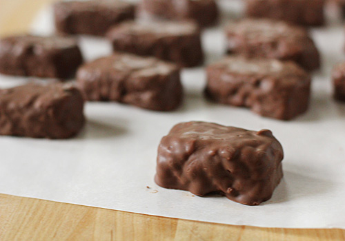 Several rectangular chocolate-covered treats rest on a sheet of parchment paper atop a wooden surface, with one treat prominently in focus in the foreground.