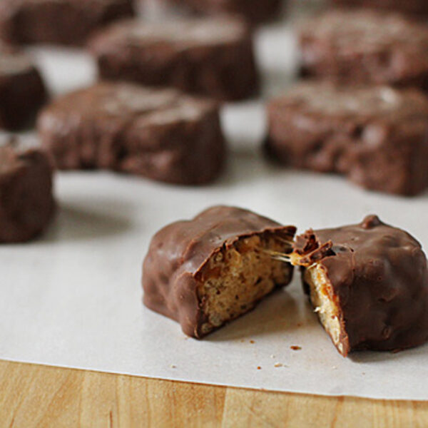 Several chocolate-covered candy bars sit on parchment paper, with one in the foreground cut in half to reveal a nougat and caramel filling inside.