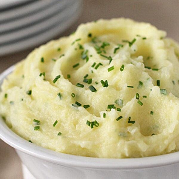 A bowl of creamy mashed potatoes garnished with chopped chives sits on a table, with a stack of white plates and a spoon in the background.