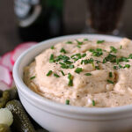 A white bowl filled with creamy dip garnished with chopped chives, surrounded by pickles, crackers, and sliced radishes, with a bottle and glass in the blurred background.