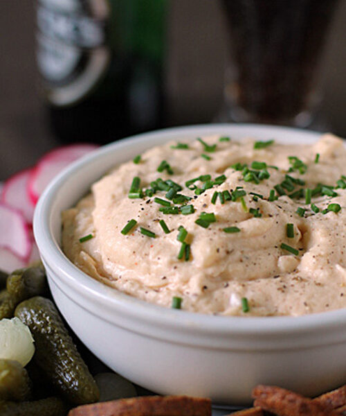 A white bowl filled with creamy dip garnished with chopped chives, surrounded by pickles, crackers, and sliced radishes, with a bottle and glass in the blurred background.