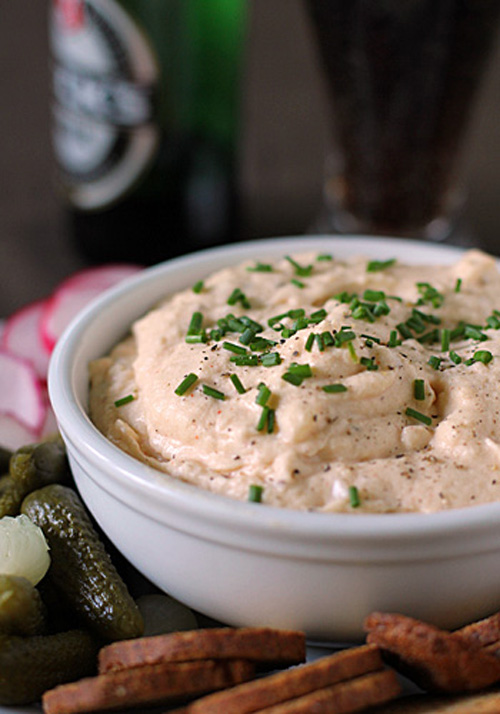 A white bowl filled with creamy dip garnished with chopped chives, surrounded by pickles, crackers, and sliced radishes, with a bottle and glass in the blurred background.