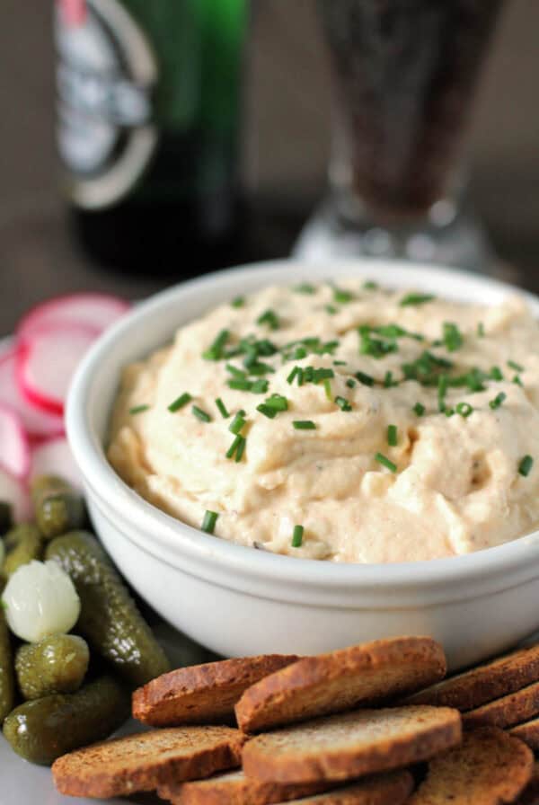 A bowl of creamy Obatzda topped with chives, served with sliced radishes, cornichons, and toasted bread rounds.