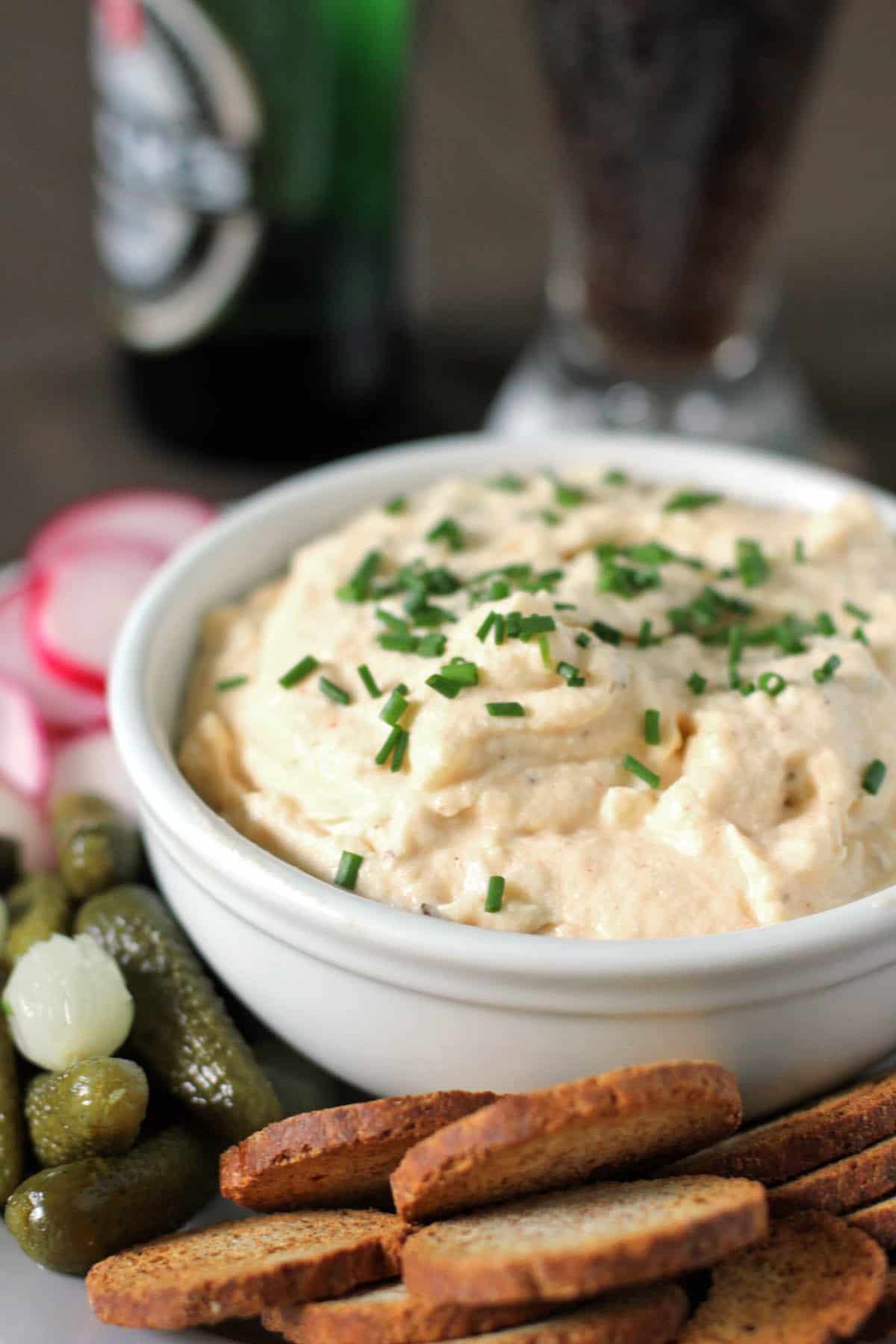 A bowl of creamy Obatzda topped with chives, served with sliced radishes, cornichons, and toasted bread rounds.