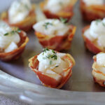 A silver tray holds several bite-sized appetizers made of crispy bacon cups filled with diced pears, creamy cheese, and garnished with fresh thyme.