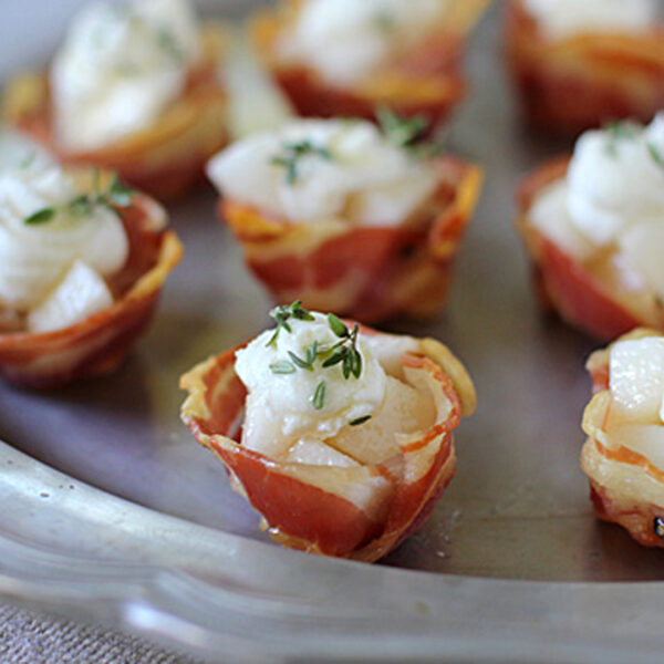 A silver tray holds several bite-sized appetizers made of crispy bacon cups filled with diced pears, creamy cheese, and garnished with fresh thyme.