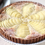 A baked pear tart with a golden crust, featuring thinly sliced pears arranged in a decorative pattern and lightly dusted with powdered sugar, placed on a white plate.