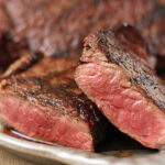 Two slices of medium-rare steak with a browned, seared exterior and a pink, juicy interior are displayed on a plate, with another piece of steak in the background.