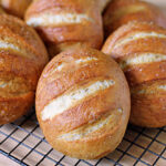 Several golden brown bread rolls with slashed tops are cooling on a black wire rack placed on a wooden surface. The rolls have a shiny, crisp crust and appear freshly baked.