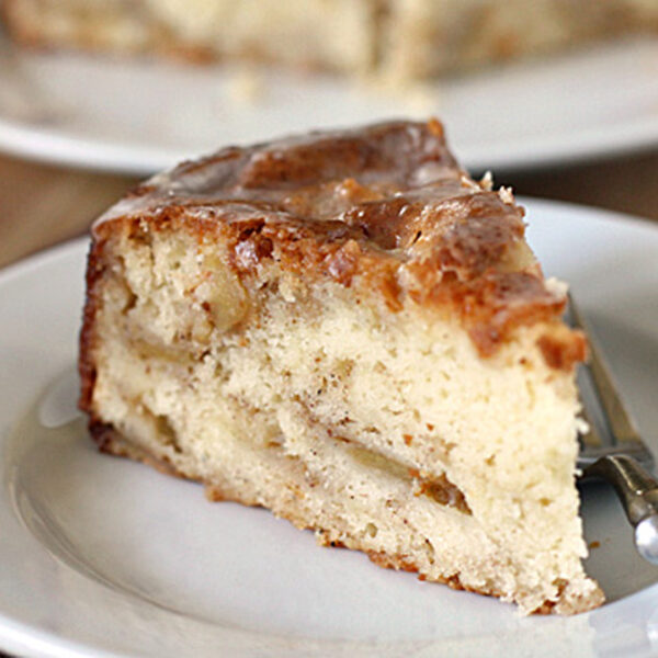 A slice of moist cake with chunks of fruit, possibly apples, sits on a white plate with a fork. The cake has a golden-brown top and a light, fluffy texture inside.