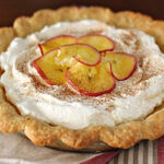 A pie with a golden, fluted crust, topped with fluffy whipped cream, cinnamon, and thin slices of apple arranged in a decorative pattern, sits on a folded napkin.