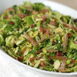 A serving dish filled with shredded Brussels sprouts mixed with crispy bacon pieces and garnished with herbs, with a serving spoon resting on the side.