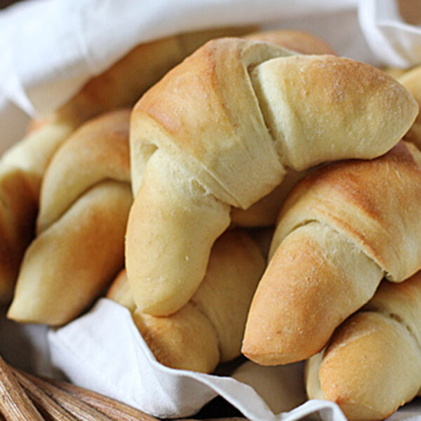 A close-up of several golden-brown crescent rolls stacked in a basket lined with a white cloth. The bread looks soft and freshly baked.