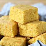 A close-up of several pieces of golden, crumbly cornbread stacked on a cloth, with a soft and moist texture visible.