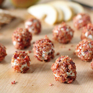 Small cheese balls coated with crispy bacon bits are arranged on a wooden surface, with sliced pears and a knife blurred in the background.