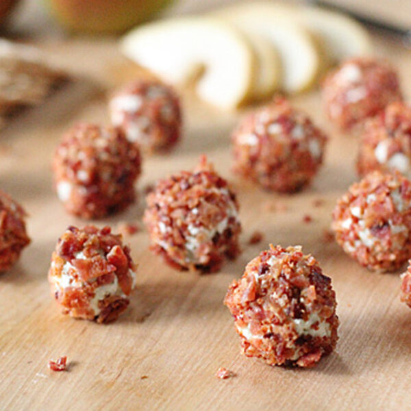 Small cheese balls coated with crispy bacon bits are arranged on a wooden surface, with sliced pears and a knife blurred in the background.