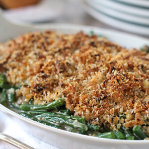 A white baking dish filled with green bean casserole, topped with a golden brown breadcrumb crust, sits on a table beside a spoon and stacked plates.