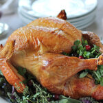 A golden-brown roasted turkey garnished with fresh herbs and greens, placed on a white platter ready to be served, with stacked plates and cranberry sauce visible in the background.