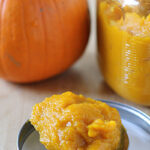 A spoonful of pumpkin puree sits on a jar lid, with a jar of more puree and a whole pumpkin in the background on a wooden surface.
