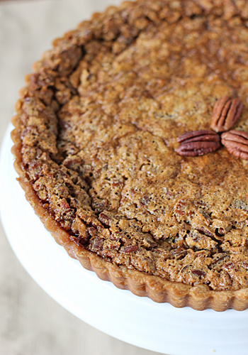 A close-up of a pecan pie with a golden, textured crust, topped with a small cluster of whole pecans in the center, displayed on a white cake stand.