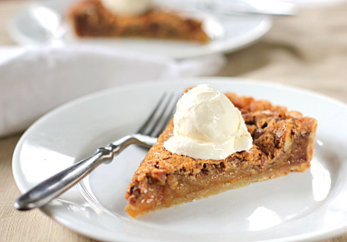 A slice of pecan pie topped with a scoop of vanilla ice cream is served on a white plate with a fork, with another plate and napkin blurred in the background.