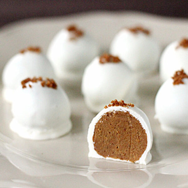 Eight white chocolate-coated truffles topped with brown crumbs are arranged on a white plate, with one truffle cut open to reveal a smooth brown filling inside.